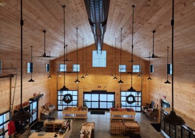 A wide-angle view of a wooden interior with high ceilings, multiple windows, and overhead lighting. The space features counters and produce displays, with wreaths hanging on the doors.