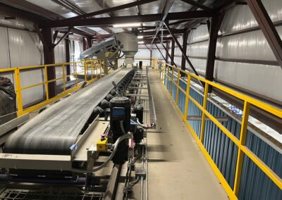 A large industrial conveyor belt system inside a metal building, with yellow railings on both sides and overhead lighting. Several pieces of machinery and control equipment are visible along the conveyor.