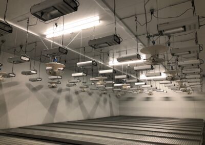 A large, empty indoor facility with multiple ceiling-mounted light fixtures and an array of parallel metal shelves on the floor.