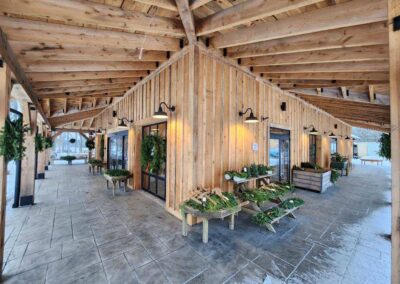 A wooden pavilion with plants displayed on benches under its roof. It is supported by timber beams and has potted evergreens positioned at the frontage. There is snow on the ground outside.